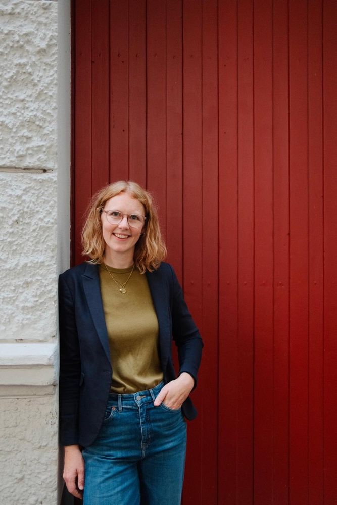 Meike Hornbostel lehnt lächelnd an einer roten Holztür, dunkler Blazer, olivfarbenes Shirt, eine Hand in der Hosentasche.